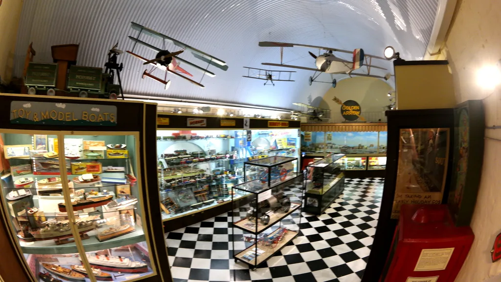 Het interieur van een speelgoedwinkel met vitrinekasten vol dozen, modelvliegtuigen aan het plafond en een zwart-wit geblokte vloer.
