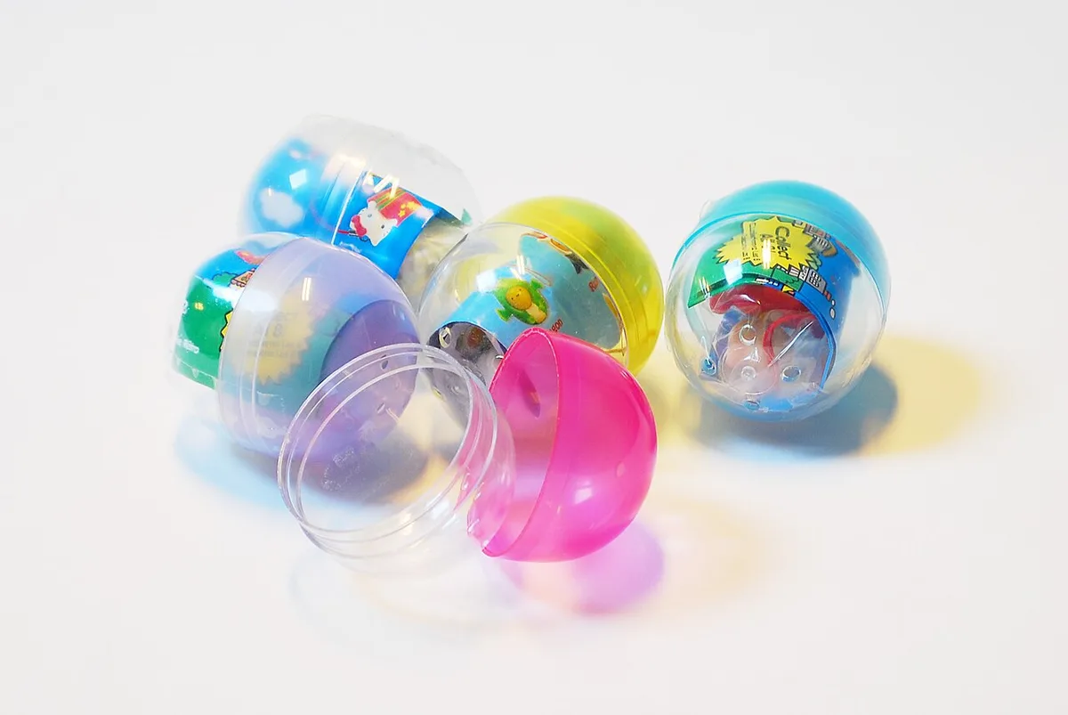 Doorzichtige plastic capsules met kleine speelgoedfiguren erin liggen op een witte ondergrond; een capsule ligt geopend in tweeën.