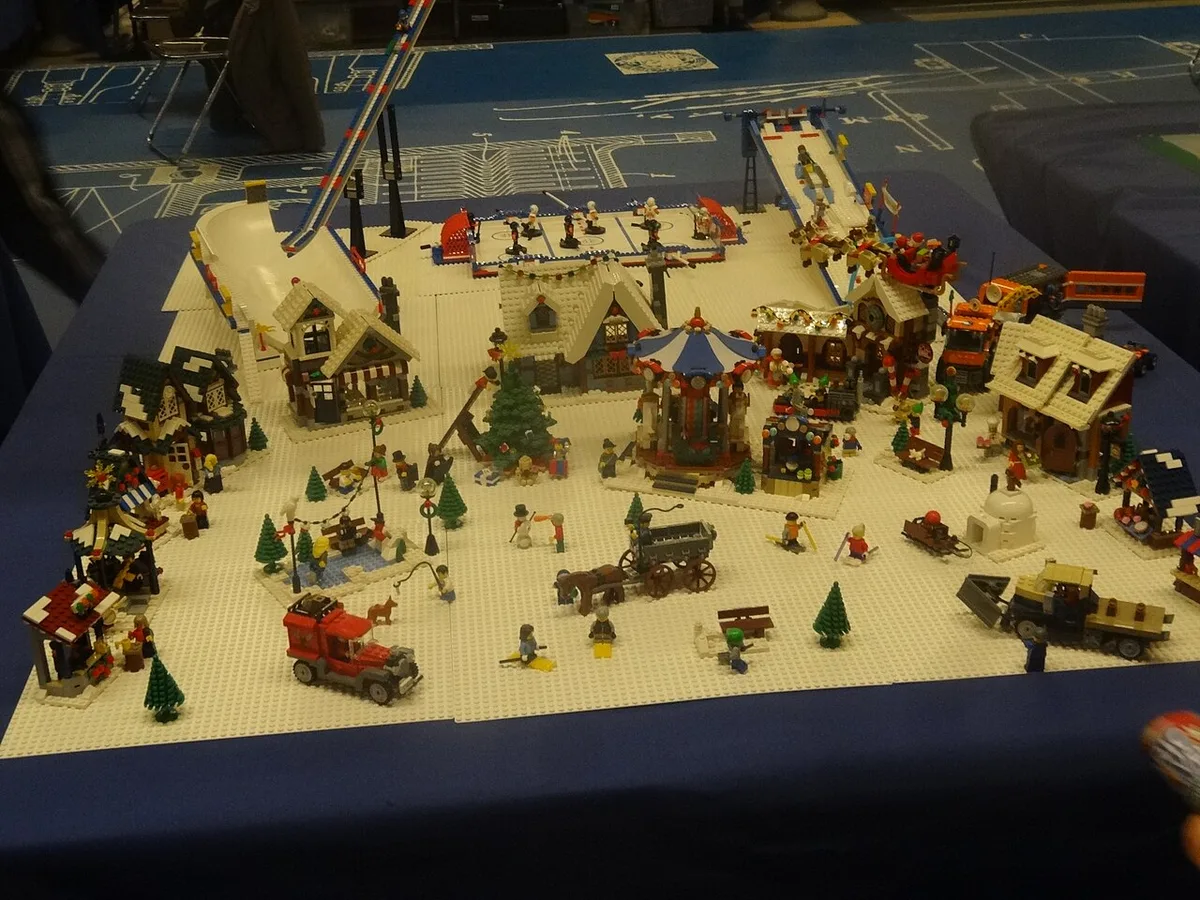Een LEGO-diorama van een besneeuwd dorp met huizen, kerstbomen, een draaimolen en minifiguren, opgesteld op een blauwe tafel.