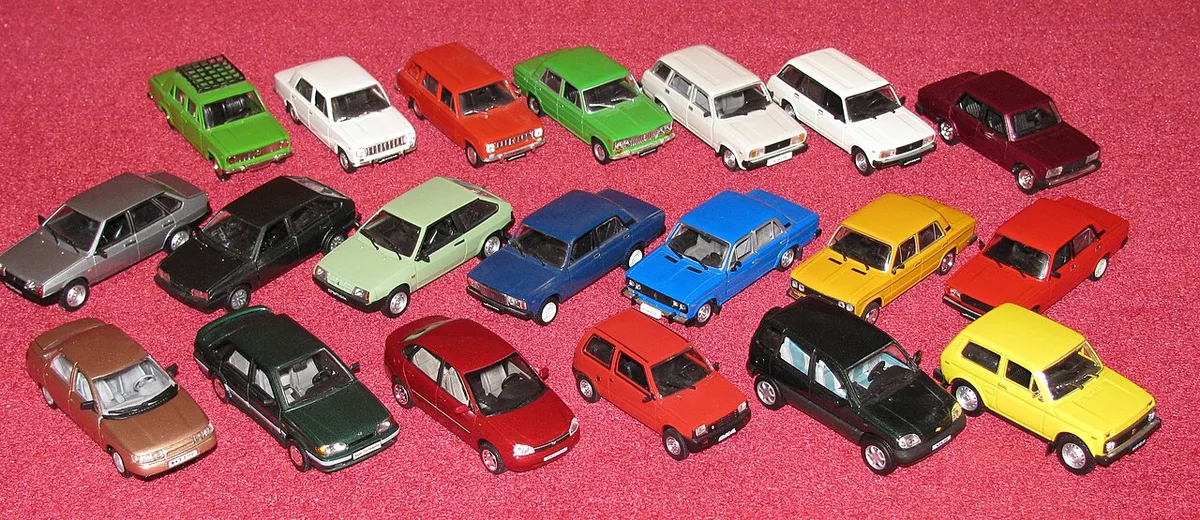 Een groep modelauto's op schaal 1:43 in uiteenlopende kleuren staat in rijen naast elkaar opgesteld op een roze ondergrond.