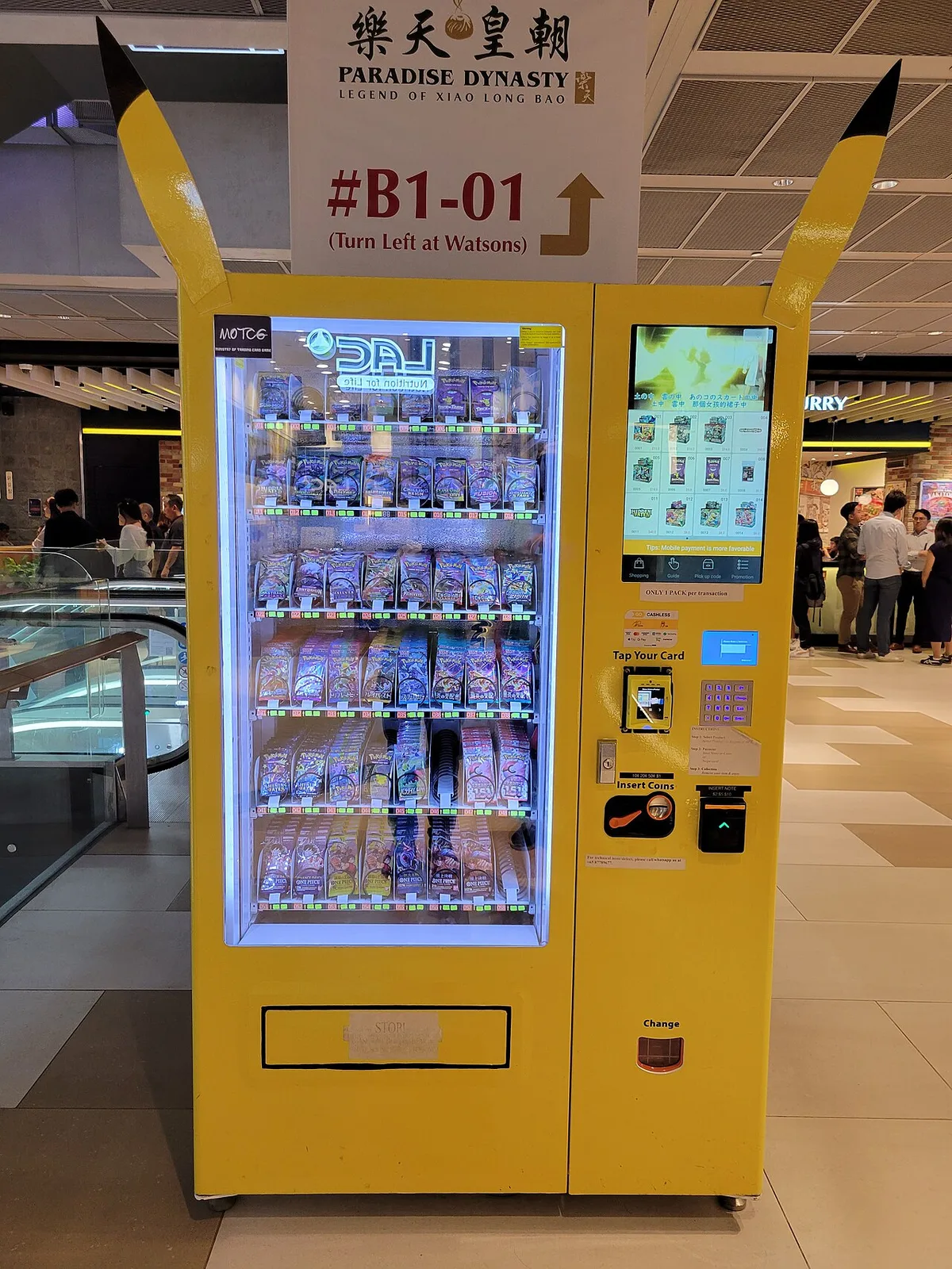 Een verkoopautomaat gevuld met pakjes Pokémonkaarten in een winkelcentrum, met bezoekers op de achtergrond.