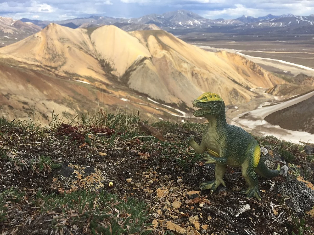 Een speelgoeddinosaurus van groen plastic staat op een met mos begroeide bergrug, met kale gele bergtoppen op de achtergrond.